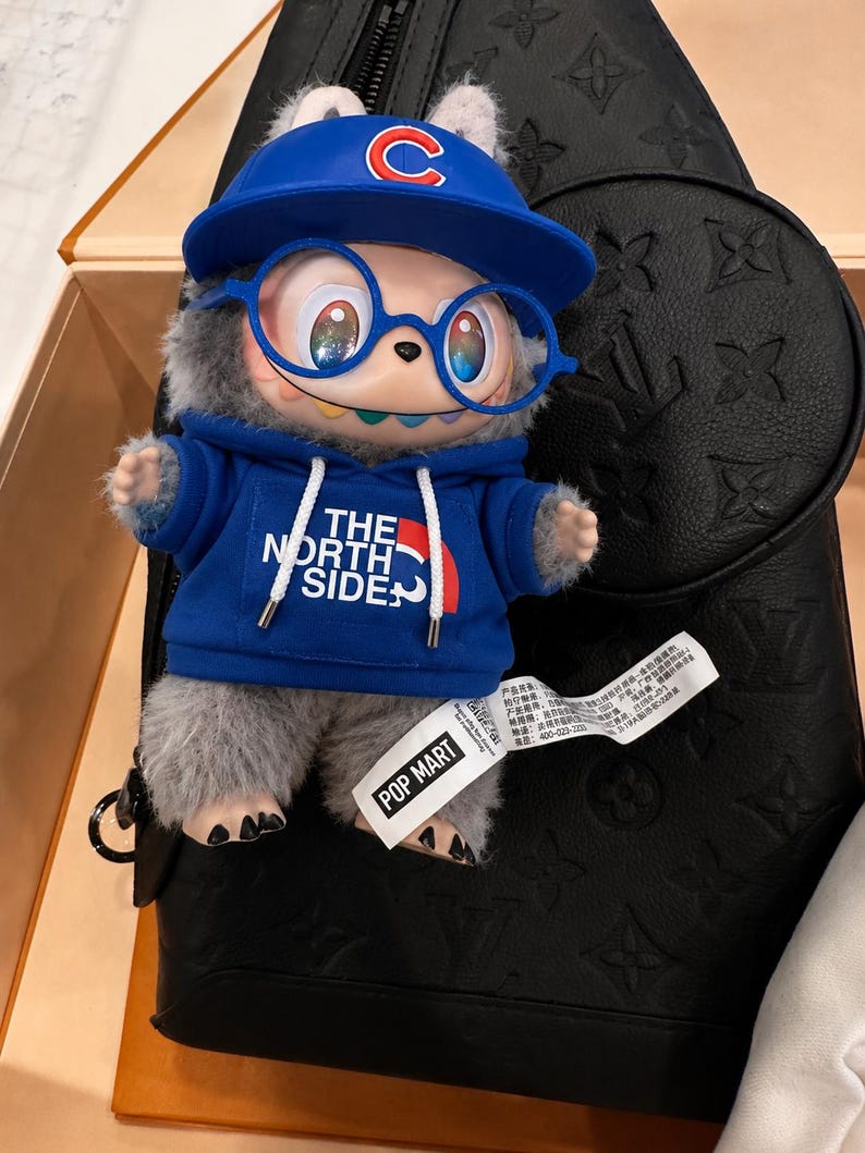 May include: A plush toy figure wearing a blue Chicago Cubs baseball cap and a blue hoodie with "THE NORTH SIDE" printed on it. The figure has blue glasses and is set against a black leather bag.