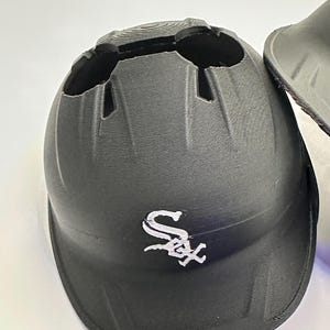 Puede incluir: Casco de b&eacute;isbol negro con el logotipo de los Chicago White Sox en blanco. El casco tiene forma redondeada con cortes en la parte superior y una visera. Fabricado con material texturizado.