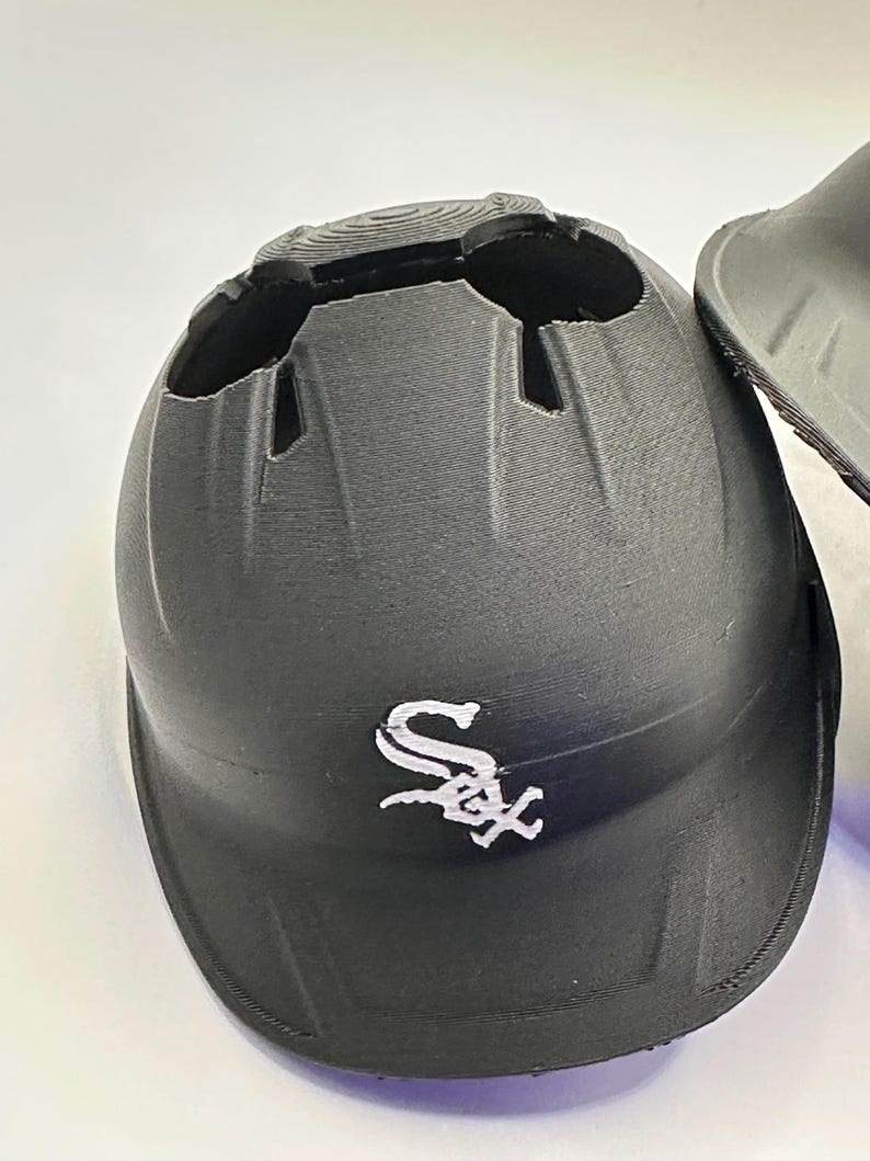Puede incluir: Casco de b&eacute;isbol negro con el logotipo de los Chicago White Sox en blanco. El casco tiene forma redondeada con cortes en la parte superior y una visera. Fabricado con material texturizado.