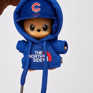 May include: A blue keychain featuring a cartoon character wearing a blue baseball cap with a white "C" logo and a matching hoodie. The hoodie has the text "THE NORTH SIDE!" and a red arc. The character has large eyes and is wearing headphones.