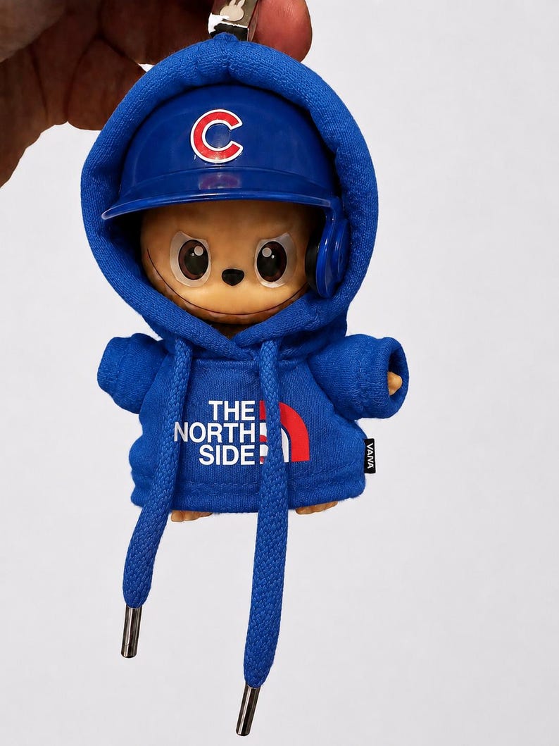 May include: A blue keychain featuring a cartoon character wearing a blue baseball cap with a white "C" logo and a matching hoodie. The hoodie has the text "THE NORTH SIDE!" and a red arc. The character has large eyes and is wearing headphones.
