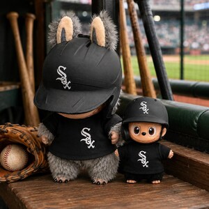 Puede incluir: Dos figuras de peluche con uniformes de b&eacute;isbol negros con el logo de los Chicago White Sox. La figura m&aacute;s grande tiene orejas peludas y un casco de b&eacute;isbol. Un guante y una pelota de b&eacute;isbol est&aacute;n en el fondo. Las figuras est&aacute;n sobre una superficie de madera.