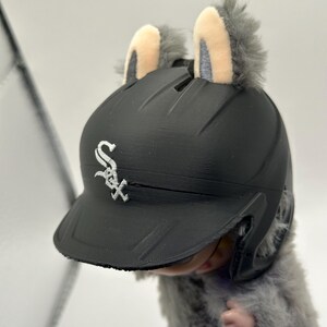 Puede incluir: Un casco de b&eacute;isbol negro con el logotipo de los Medias Blancas de Chicago, adornado con orejas grises de felpa. El casco lo lleva una peque&ntilde;a criatura peluda. Las orejas tienen un interior de color beige claro.