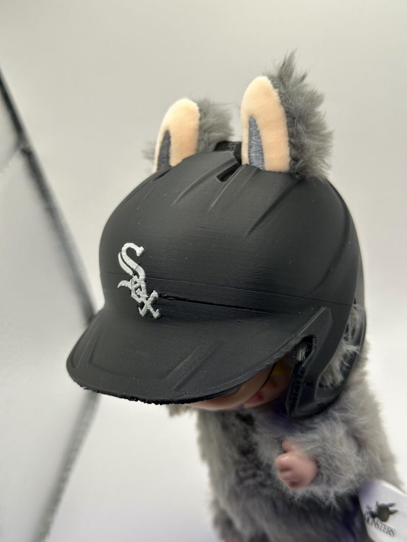 Puede incluir: Un casco de b&eacute;isbol negro con el logotipo de los Medias Blancas de Chicago, adornado con orejas grises de felpa. El casco lo lleva una peque&ntilde;a criatura peluda. Las orejas tienen un interior de color beige claro.
