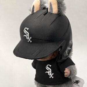 Puede incluir: Un juguete de peluche que lleva un casco y una camiseta de b&eacute;isbol negros con el logotipo de los Medias Blancas de Chicago. El juguete tiene pelo gris y orejas peque&ntilde;as. El casco y la camiseta muestran el logotipo "Sox" del equipo en blanco.