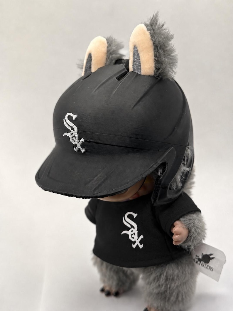 Puede incluir: Un juguete de peluche que lleva un casco y una camiseta de b&eacute;isbol negros con el logotipo de los Medias Blancas de Chicago. El juguete tiene pelo gris y orejas peque&ntilde;as. El casco y la camiseta muestran el logotipo "Sox" del equipo en blanco.