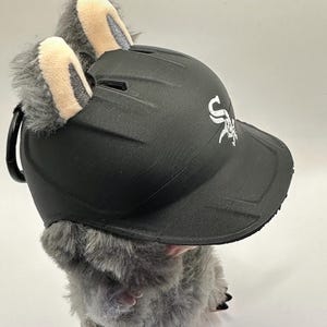 Puede incluir: Una gorra de b&eacute;isbol negra con el logotipo de los White Sox, adornada con pelaje de felpa gris y orejas forradas en beige. La gorra es parte de un art&iacute;culo novedoso, probablemente un disfraz o accesorio.