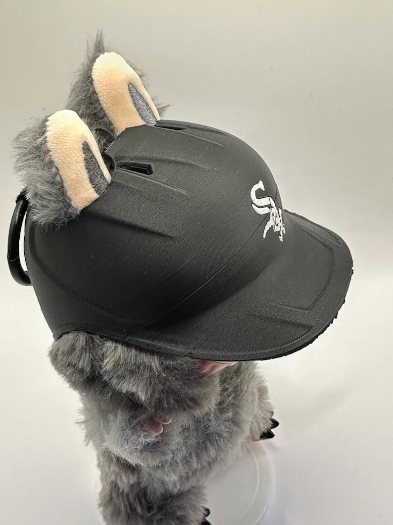 Puede incluir: Una gorra de b&eacute;isbol negra con el logotipo de los White Sox, adornada con pelaje de felpa gris y orejas forradas en beige. La gorra es parte de un art&iacute;culo novedoso, probablemente un disfraz o accesorio.
