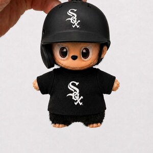 Puede incluir: Una figura negra con tem&aacute;tica de b&eacute;isbol que lleva un casco y una camiseta negros con el logotipo de los White Sox en blanco. La figura tiene una cara de dibujos animados y est&aacute; unida a un llavero plateado.