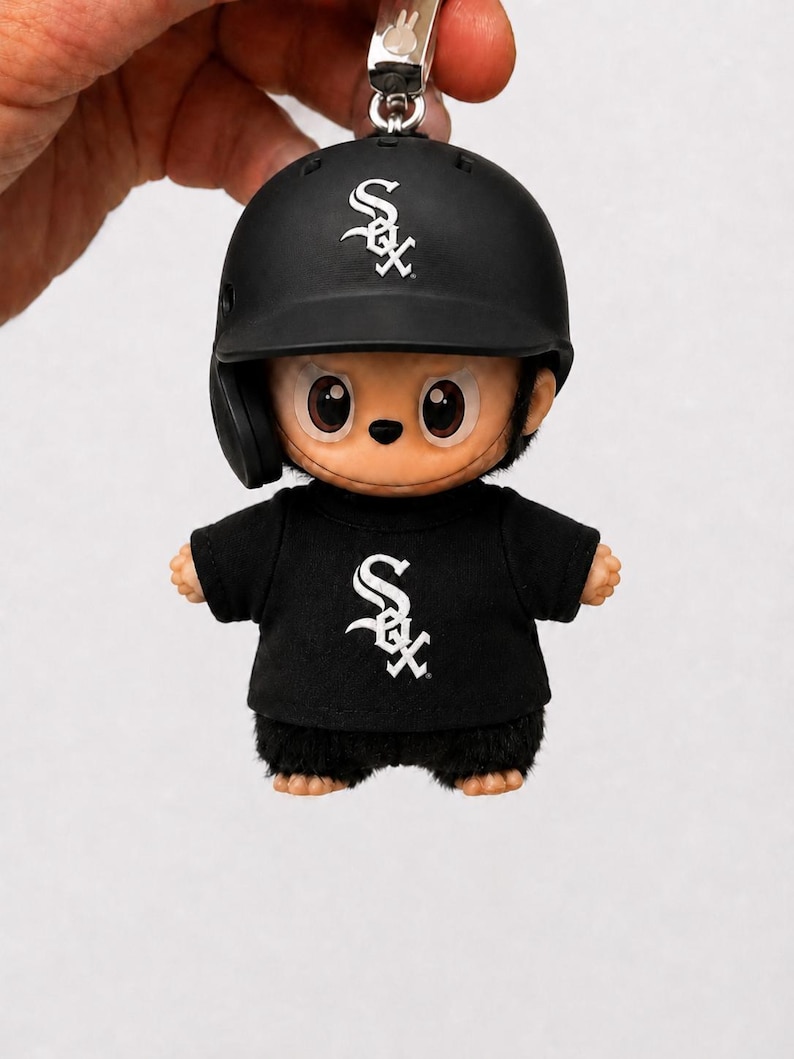 Puede incluir: Una figura negra con tem&aacute;tica de b&eacute;isbol que lleva un casco y una camiseta negros con el logotipo de los White Sox en blanco. La figura tiene una cara de dibujos animados y est&aacute; unida a un llavero plateado.