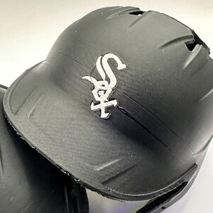 Puede incluir: Casco de b&eacute;isbol negro con el logotipo blanco de los Chicago White Sox. El casco tiene un acabado mate y una visera curva. El logotipo es una "S" y una "X" estilizadas con una cruz.