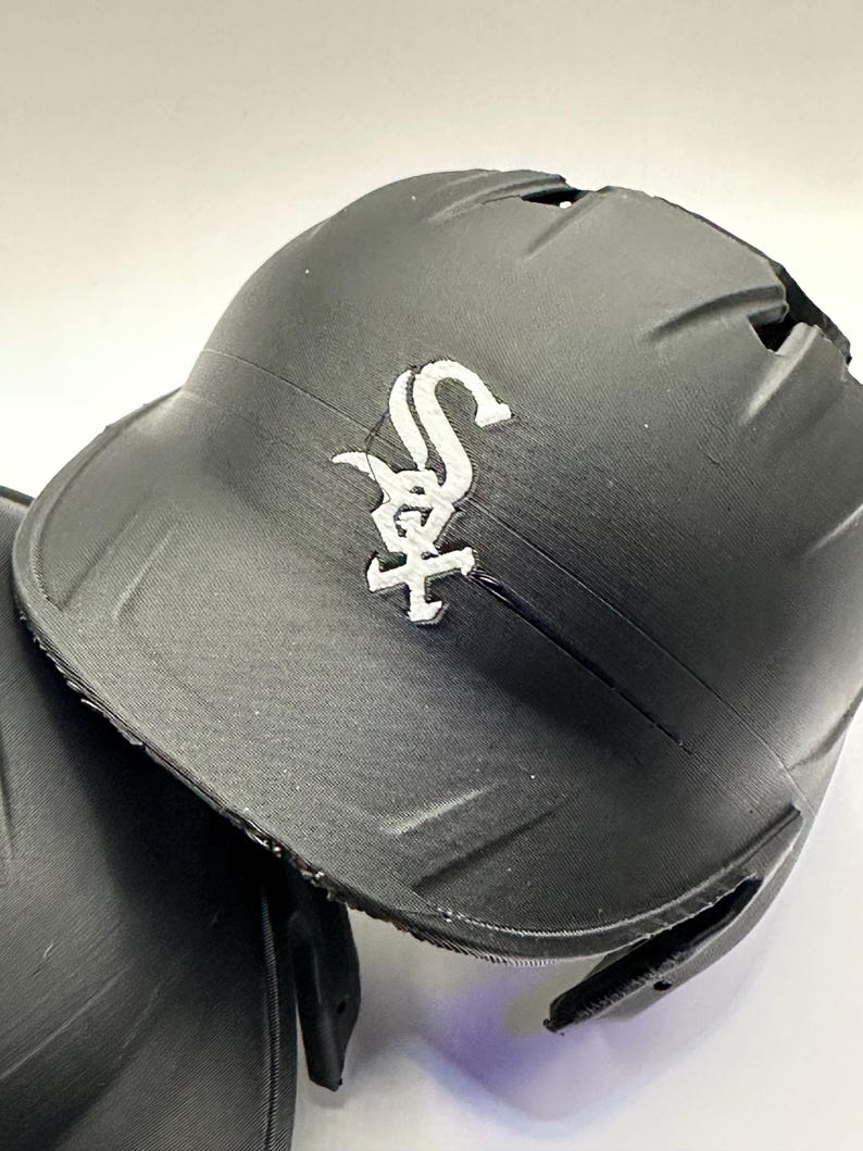 Puede incluir: Casco de b&eacute;isbol negro con el logotipo blanco de los Chicago White Sox. El casco tiene un acabado mate y una visera curva. El logotipo es una "S" y una "X" estilizadas con una cruz.