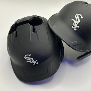 Puede incluir: Dos cascos de b&eacute;isbol negros con el logotipo de los Chicago White Sox en blanco. Un casco tiene una gran abertura en la parte superior. El otro casco est&aacute; intacto. Ambos cascos est&aacute;n sobre una superficie blanca.