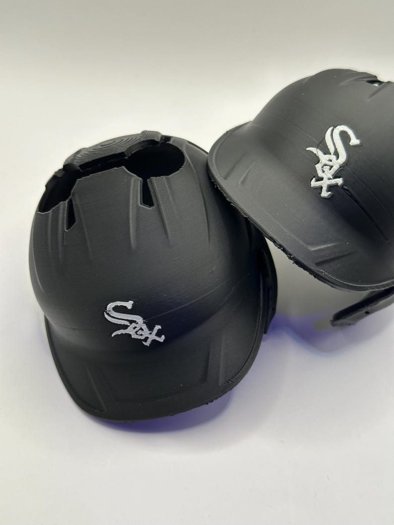Puede incluir: Dos cascos de b&eacute;isbol negros con el logotipo de los Chicago White Sox en blanco. Un casco tiene una gran abertura en la parte superior. El otro casco est&aacute; intacto. Ambos cascos est&aacute;n sobre una superficie blanca.