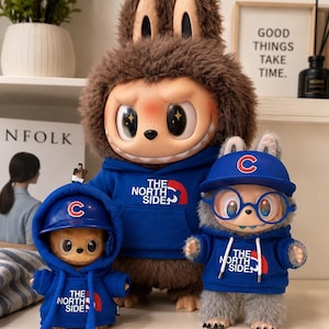 May include: Three plush figures wearing blue hoodies and baseball caps. The hoodies have "THE NORTH SIDE" printed on them. The largest figure has a furry brown head and ears. The caps have a "C" logo.