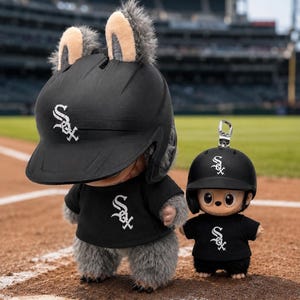 Puede incluir: Dos figuras de peluche con cascos y camisetas de b&eacute;isbol negros con el logotipo de los White Sox de Chicago. La figura m&aacute;s grande tiene orejas peludas en el casco. Est&aacute;n en un campo de b&eacute;isbol.