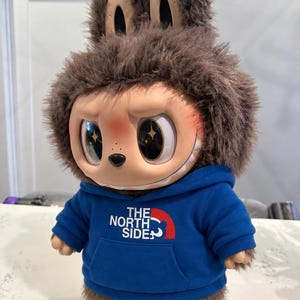 May include: A plush doll with a brown furry head and ears, wearing a blue hoodie with "THE NORTH SIDE" printed on it. The doll has large, expressive eyes with star-shaped reflections and a smiling face.