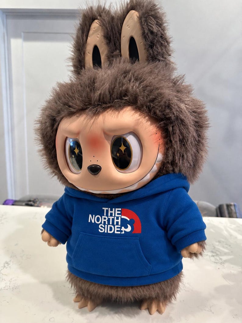 May include: A plush doll with a brown furry head and ears, wearing a blue hoodie with "THE NORTH SIDE" printed on it. The doll has large, expressive eyes with star-shaped reflections and a smiling face.