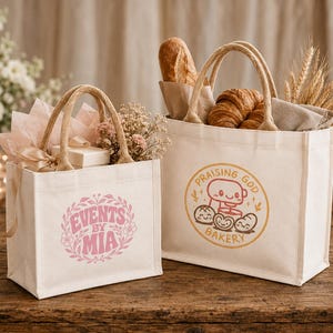 May include: Two off-white canvas tote bags with jute handles. The smaller bag features a pink floral wreath design with the text "EVENTS BY MIA". The larger bag displays an orange circular logo with a mixer and bread, reading "PRAISING GOD BAKERY".