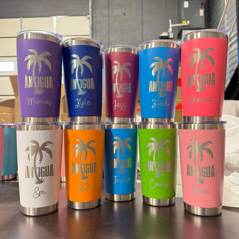 May include: A collection of insulated tumblers in various colors, including purple, blue, red, and pink. Each tumbler features a silver palm tree design and the word "Antigua" with a name below. The tumblers are stainless steel and have a silver rim.