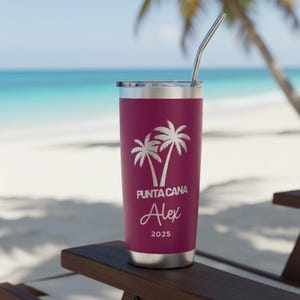 May include: A maroon stainless steel tumbler with a silver lid and straw. The tumbler features a white graphic of two palm trees and the text "PUNTA CANA" above the name "Alex" and the year "2025". The tumbler is sitting on a wooden chair with a beach and ocean in the background.