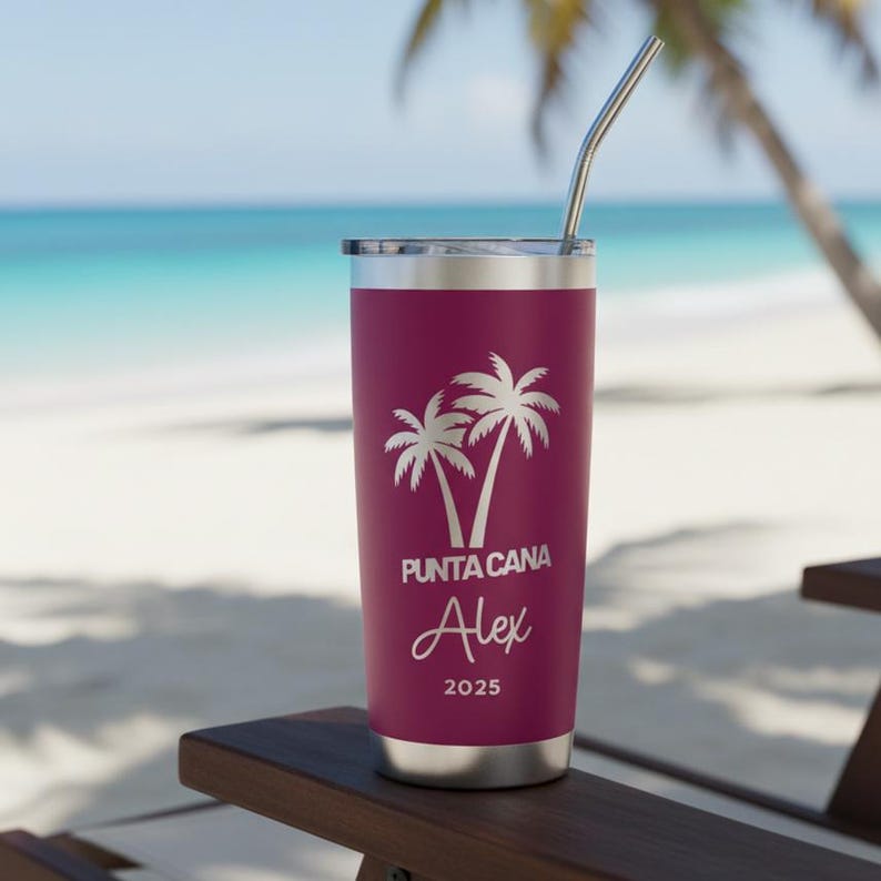 May include: A maroon stainless steel tumbler with a silver lid and straw. The tumbler features a white graphic of two palm trees and the text "PUNTA CANA" above the name "Alex" and the year "2025". The tumbler is sitting on a wooden chair with a beach and ocean in the background.