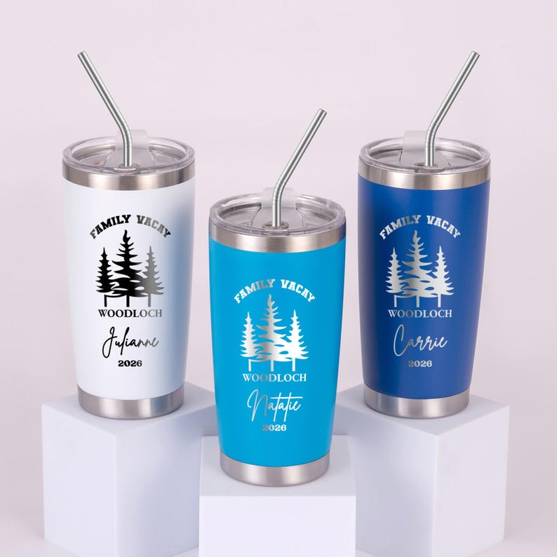Puede incluir: Tres vasos de acero inoxidable con tapas y pajitas. Uno es blanco, otro turquesa y otro azul. Cada vaso tiene un gr&aacute;fico plateado de &aacute;rboles y las palabras "Family Vacay" y "Woodloch". Cada uno tiene un nombre y el a&ntilde;o 2026.