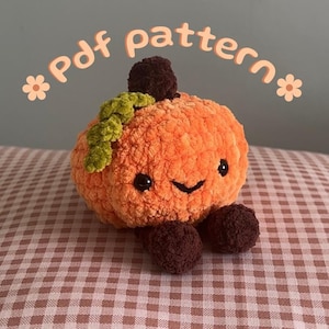 Puede incluir: Un peluche de calabaza tejido a ganchillo con hilo naranja, un tallo marrón oscuro, pies marrón oscuro y una hoja verde texturizada. El peluche tiene ojos de seguridad negros y una sonrisa bordada. El texto "Pdf pattern" aparece arriba.