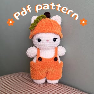 May include: A white crocheted plush toy wearing an orange pumpkin-shaped hat with a green vine detail and orange overalls with two dark brown buttons. The text "Pdf pattern" is displayed in orange above the plush toy with orange flower icons.