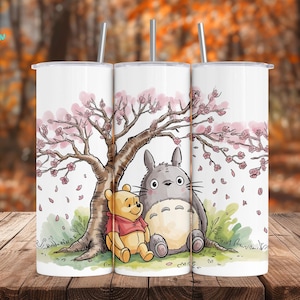 Peut inclure: Gobelet blanc avec une illustration de dessin animé de Winnie l'ourson et Totoro sous un cerisier en fleurs. Le design comprend des fleurs roses et un tronc d'arbre brun. Le gobelet a une paille argentée.