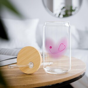 May include: A clear glass can-shaped drinking glass with a pink stethoscope design. The glass has a bamboo lid and a clear straw. The glass is sitting on a wooden table.