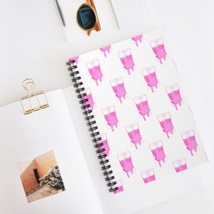 Peut inclure: Un cahier à spirales avec une couverture blanche ornée d'un motif répété de poches de perfusion roses. Le cahier est ouvert, révélant une photographie sur la page de gauche. Un trombone doré et des lunettes de soleil sont également visibles.