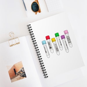 Peut inclure: Un carnet à spirales blanc ouvert, orné d'un dessin de six tubes à essai avec des bouchons colorés. Les bouchons sont bleus, rouges, jaunes, verts, violets et roses. Le carnet est posé sur une surface blanche.