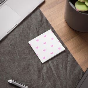 Peut inclure: Un bloc-notes carré blanc avec un motif de petits cœurs roses. Les notes sont posées sur une surface gris foncé, à côté d'un ordinateur portable et d'un stylo. Une plante en pot est en arrière-plan.