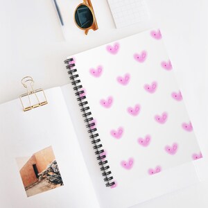 May include: White spiral-bound notebook with a pattern of pink heart-shaped faces. The notebook is open, revealing blank pages and a photo. A gold binder clip, sunglasses, and a pen are also visible.