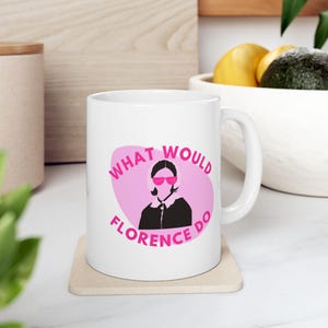 Peut inclure: Mug en céramique blanche avec l'inscription rose "WHAT WOULD FLORENCE DO", entourant une silhouette de femme portant des lunettes de soleil roses. Le mug est posé sur un dessous de verre beige.