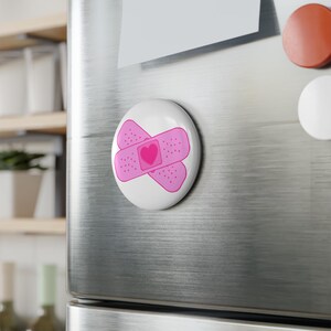 Peut inclure: Un aimant ovale blanc avec un motif de pansement rose et un cœur au centre. Le pansement est croisé. L'aimant est fixé à une porte de réfrigérateur en acier inoxydable.