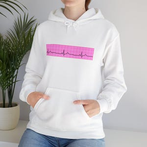 Peut inclure: Un sweat à capuche blanc avec une poche avant et une capuche à cordon. Le sweat à capuche présente un motif rectangulaire rose avec un design d'électrocardiogramme noir. Les manches sont longues et la matière semble être un tissu doux.
