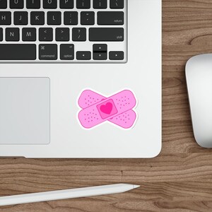 Peut inclure: Un autocollant rose représentant deux pansements qui se chevauchent avec un cœur au centre. Les pansements ont de petits détails pointillés. L'autocollant est sur une surface blanche, près d'un clavier d'ordinateur portable et d'une table en bois.