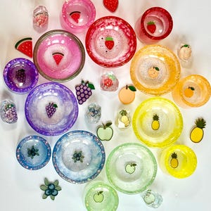 May include: A collection of colorful resin bowls and fruit-themed embellishments. The bowls feature embedded glitter and fruit slices, including watermelon, strawberries, grapes, blueberries, apples, oranges, and pineapples. The bowls are in various sizes and colors.