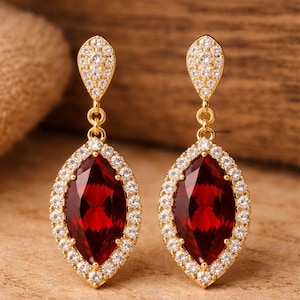May include: A pair of gold-toned earrings featuring large, marquise-cut, deep red gemstones. Each gemstone is surrounded by a halo of small, clear crystals. The earrings have a teardrop-shaped top with more crystals.