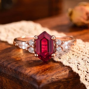 May include: A rose gold ring featuring a large, faceted, hexagonal ruby. The ring is accented with smaller white stones and set on a wooden surface with a lace ribbon.