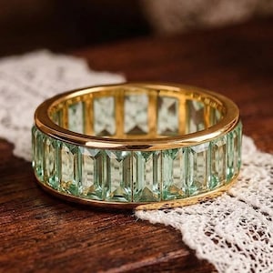 Op de afbeelding: Een goudkleurige ring met een band van rechthoekige, lichtgroene edelstenen. De ring ligt op een houten oppervlak met een wit kanten detail. De edelstenen zijn rond de band gerangschikt.