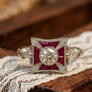 May include: A vintage-style ring with a central round diamond, surrounded by square red gemstones and smaller diamonds. The ring is set in a silver-colored metal and rests on a wooden surface with a lace overlay. The ring's design is symmetrical and ornate.