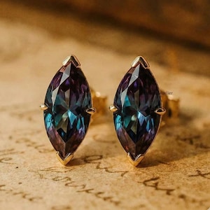 May include: A pair of gold stud earrings featuring marquise-cut gemstones that change colour. The gemstones transition from teal to purple, set in a gold bezel. The earrings are displayed on a vintage paper background.
