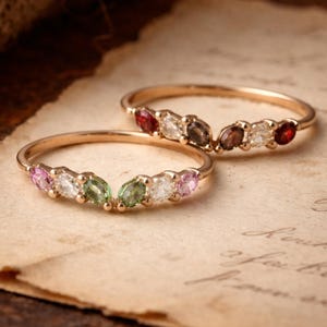 Peut inclure: Deux bagues en or ornées de pierres précieuses colorées. L'une présente des pierres roses, vertes et blanches, l'autre des pierres rouges, marron et blanches. Posées sur un papier vieilli, elles évoquent le vintage.