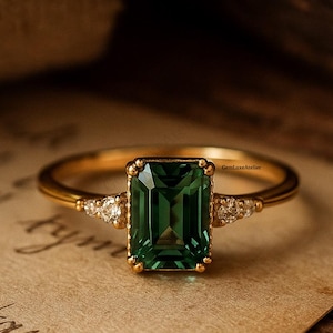 Könnte beinhalten: Ein goldener Ring mit einem rechteckigen, smaragdgeschliffenen grünen Edelstein als Mittelpunkt. Kleine, dreieckige Akzentsteine flankieren den Hauptstein. Der Ring befindet sich auf einem Papierhintergrund im Vintage-Stil.