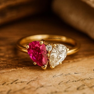 以下が含まれることがあります： ゴールドのリング。鮮やかなピンク色のペアシェイプの宝石と、透明なペアシェイプの宝石が特徴です。2つの石の間には小さなダイヤモンドがセットされています。リングは、質感のある明るい茶色の表面に置かれています。GemLuxeAtelier.