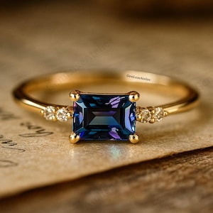 May include: A gold ring featuring a rectangular, deep blue-purple gemstone. Small, clear stones are set either side of the main stone. The ring is displayed on a textured, light brown surface.