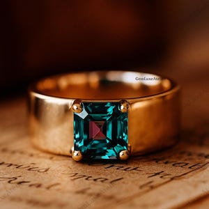 Puede incluir: Un anillo de oro con una gran piedra preciosa de color verde azulado de corte cuadrado con un toque de púrpura en el centro. El anillo tiene una banda gruesa y pequeños detalles dorados. El anillo está sobre un fondo de papel vintage.